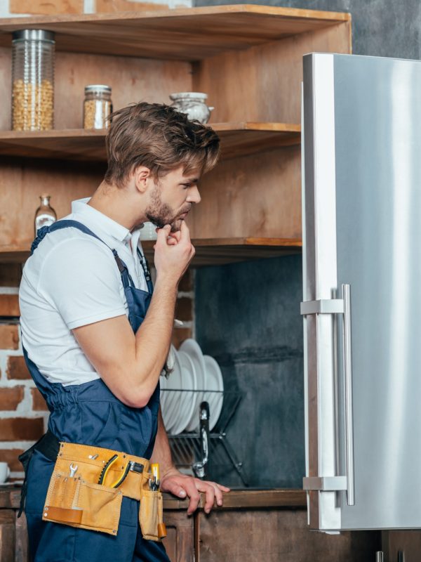 young repairman with tool belt looking at broken refrigerator